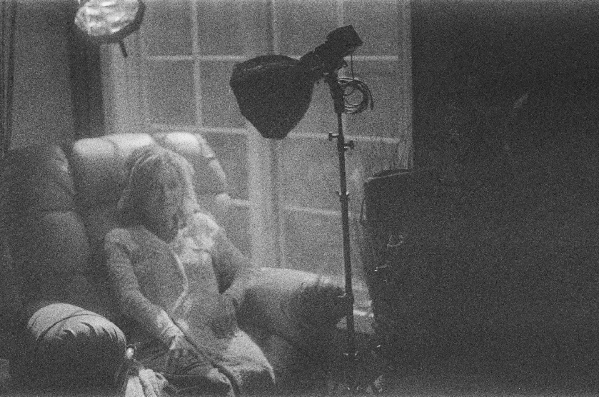 Black and white portrait of a performer in a chair beside studio lighting, seen through a window